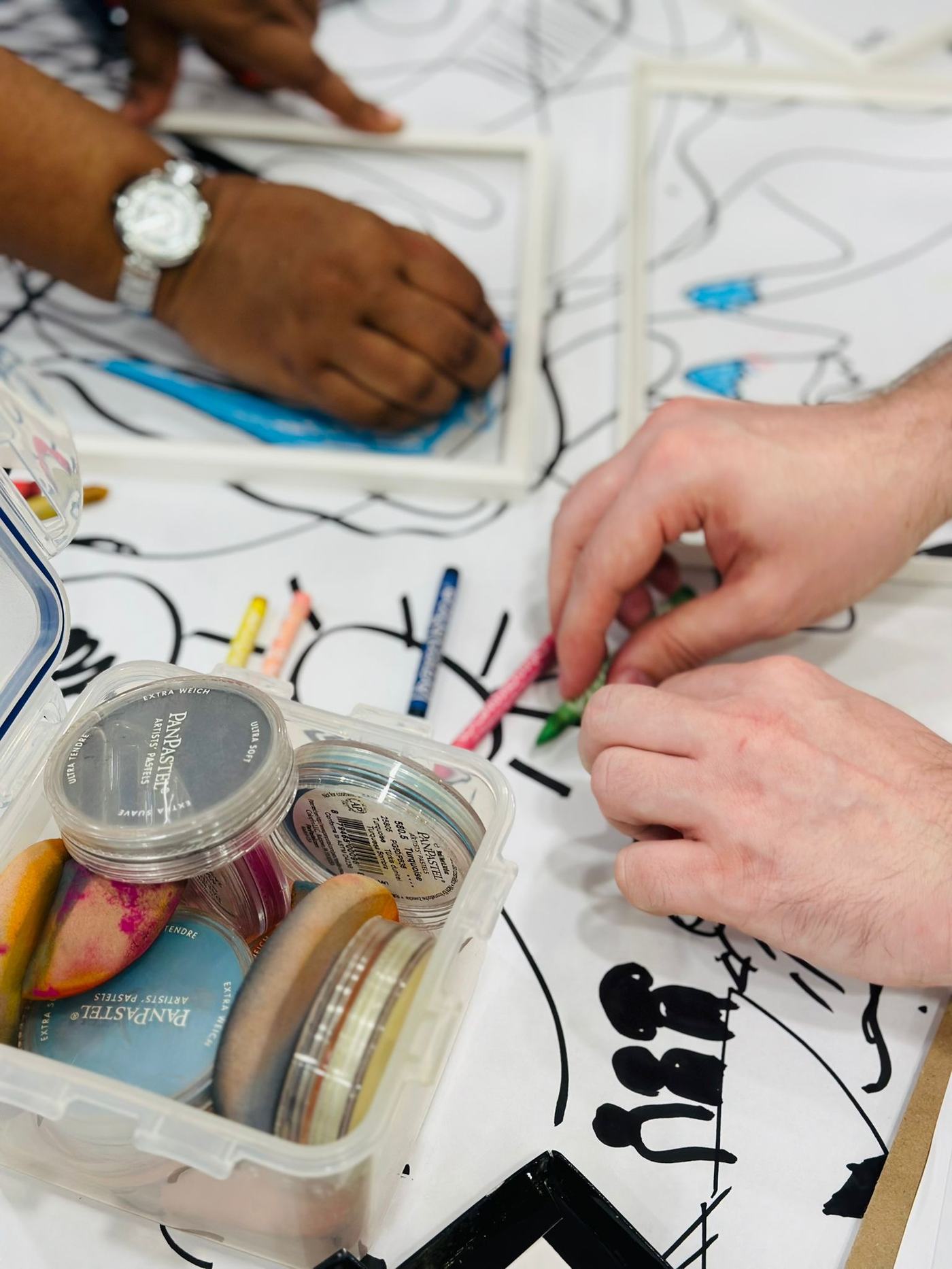 Two people choosing pastels and drawing over a large shared line drawing at a workshop